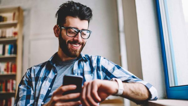 A business-casual dressed man holding a phone and glancing at his smart watch.